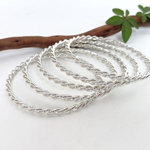Puede incluir: Un conjunto de cinco brazaletes de color plateado, cada uno con una textura trenzada similar a una cuerda. Las pulseras circulares están dispuestas en una pila ligeramente superpuesta, mostrando un acabado metálico pulido y brillante.