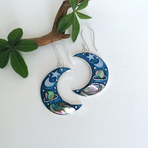 May include: A pair of crescent moon earrings. The earrings have a dark blue enamel finish with silver details, including stars and a planet. The lower part of the moon features an iridescent abalone shell inlay.
