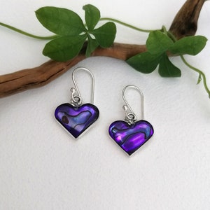 May include: A pair of silver heart-shaped earrings with purple abalone shell inlays. The earrings have a simple hook fastening.