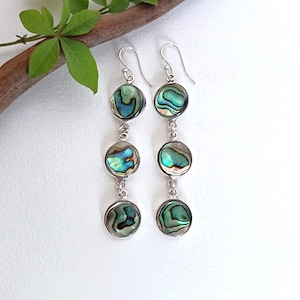 May include: A pair of drop earrings featuring three circular abalone shell inlays set in silver-toned metal. Each earring consists of three vertically linked discs displaying iridescent green, blue, and brown patterns, suspended from a silver hook.