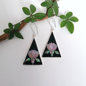 Può includere: Un paio di orecchini color argento con base triangolare nera. Ogni orecchino presenta un motivo floreale rosa con accenti verdi e blu iridescenti. Gli orecchini sono appesi a ganci d'argento.