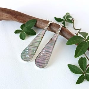 May include: A pair of long, teardrop-shaped earrings featuring iridescent, wavy-textured abalone shell inlays set within polished silver-toned metal frames. The earrings have a smooth, reflective surface that displays shifting hues of pink, green, and grey.