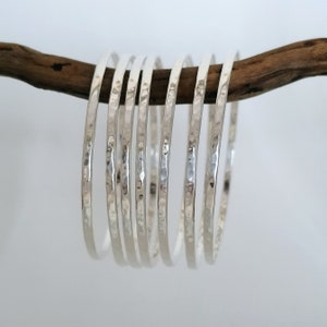 May include: A set of ten silver hammered bangle bracelets. The bracelets are stacked on top of each other and are resting on a piece of driftwood.