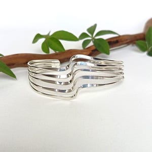 Può includere: Un bracciale a fascia in argento composto da cinque bande metalliche parallele e lucide che si curvano in un motivo a onde. Il design aperto è costituito da fili d'argento lisci e riflettenti che mantengono una forma uniforme.