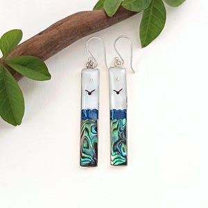 May include: A pair of rectangular drop earrings featuring a white top section with a small black clock hand graphic, a thin blue horizontal band, and a lower section of iridescent abalone shell. The earrings hang from silver-toned metal hooks.