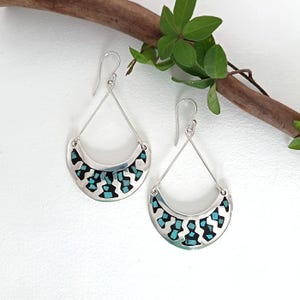 May include: A pair of silver earrings with a crescent-shaped design. The earrings feature a turquoise and black mosaic pattern within the crescent. The earrings hang from a silver wire and are displayed on a white background with a branch and green leaves.