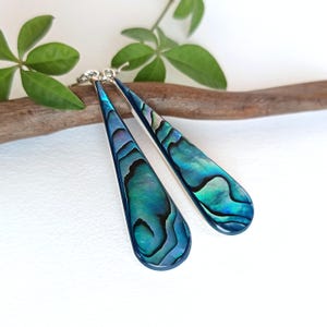 Könnte beinhalten: Ein Paar tropfenförmige Ohrringe mit schillernden blau-grünen Abalone-Muscheleinlagen. Die Ohrringe haben eine silberfarbene Metalleinfassung und werden vor einem weißen Hintergrund mit einem Ast und grünen Blättern präsentiert.