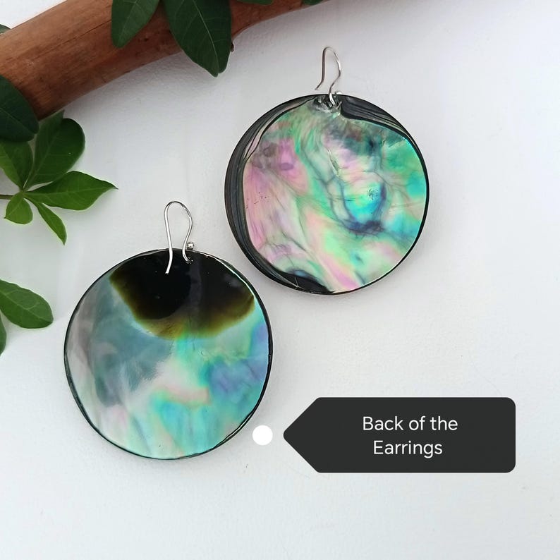 May include: Two iridescent, round earrings with silver hooks. The earrings have a black border and display a swirling pattern of green, blue, and pink hues. The image is taken from above, showing the back of the earrings.