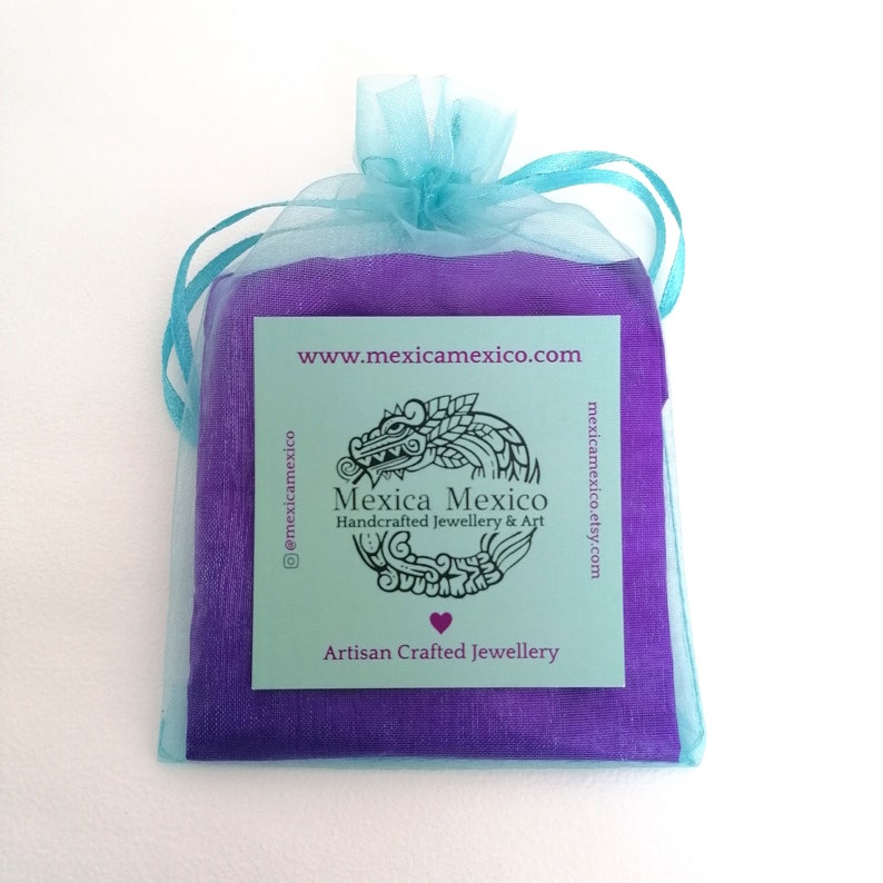 May include: A purple pouch with a turquoise drawstring, containing a small card with the text "Mexica Mexico Handcrafted Jewellery & Art" and the website address "www.mexicamexico.com". The card also features a stylized image of a serpent and the text "Artisan Crafted Jewellery".
