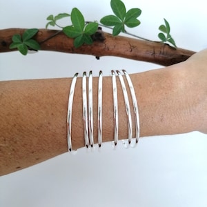 Op de afbeelding: Een set van zes zilveren armbandjes gestapeld op de pols van een persoon. De armbandjes hebben allemaal dezelfde breedte en een gladde, gepolijste afwerking.