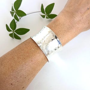 Può includere: Un ampio bracciale a fascia in argento lucido caratterizzato da una distinta texture martellata su tutta la superficie. La fascia metallica presenta una forma leggermente concava e curva che avvolge il polso, riflettendo la luce brillante.