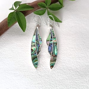 May include: A pair of long, slender drop earrings featuring iridescent abalone shell inlays set within silver-toned metal frames. The earrings have a curved, leaf-like shape with a segmented design and silver hook-style ear wires for attachment.