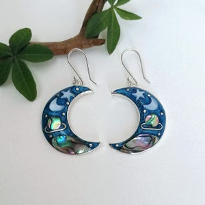 May include: A pair of crescent moon earrings with a celestial design. The earrings feature a dark blue enamel base with white stars, a moon, and a colorful abalone shell inlay. The earrings have silver-colored ear wires.