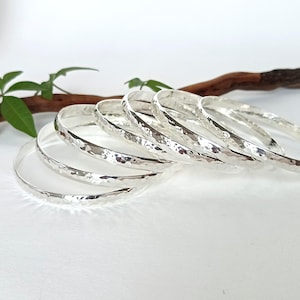 May include: A set of six silver-colored bangles arranged in a stack. Each bangle features a hammered, textured surface that reflects light, creating a bright, metallic appearance. The circular bracelets are positioned in a cascading formation.