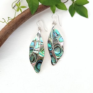 Puede incluir: Un par de pendientes colgantes con forma de hoja, incrustados con nácar de abulón iridiscente. La concha muestra patrones en espiral con tonos azules, verdes y morados, engastados en bordes de metal plateado con ganchos curvos para las orejas.