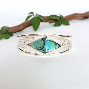Puede incluir: Una pulsera brazalete de plata con una incrustación central en forma de diamante de concha de abulón iridiscente. La banda de metal tiene un acabado liso con detalles de alambre trenzado en los lados donde la banda se une al engaste.