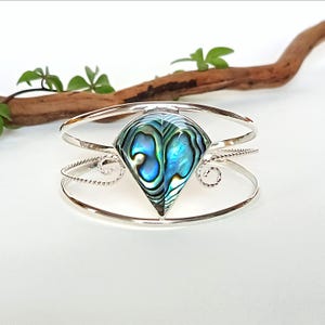 Könnte beinhalten: Ein silbernes Armreif-Armband mit einem birnenförmigen Mittelstück aus Abalone-Muschel mit schillernden blau-grünen Farbtönen. Das Armband hat ein dreibändiges Design mit dekorativen gedrehten Drähten und Wirbelakzenten.