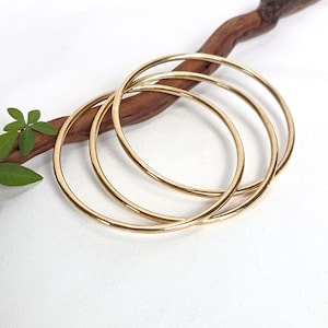 May include: Three thin, polished gold-tone bangle bracelets with a smooth, circular design. The bangles are stacked together, showcasing a reflective metallic finish. The jewellery is presented with a brown wooden branch and green leaves.