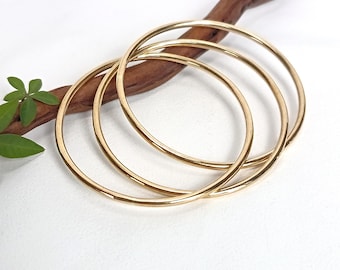 May include: Three thin, polished gold-tone bangle bracelets with a smooth, circular design. The bangles are stacked together, showcasing a reflective metallic finish. The jewelry is presented with a brown wooden branch and green leaves.