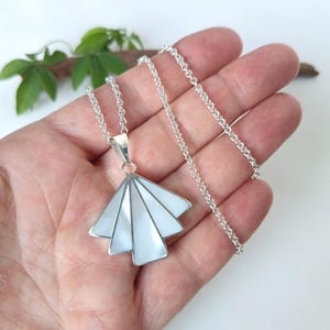 Peut inclure: Un collier en argent avec un pendentif en forme d'éventail incrusté de segments de nacre blanche. Le pendentif est suspendu à une fine chaîne en argent au fini poli, présentant un design géométrique aux lignes épurées et aux bords métalliques.