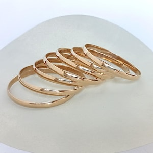 Può includere: Un set di sei bracciali rigidi in metallo color oro rosa lucido. Ogni bracciale presenta un design a fascia circolare liscia, arrotondata e continua con una superficie riflettente che cattura la luce, creando un aspetto uniforme e semplice per i gioielli.