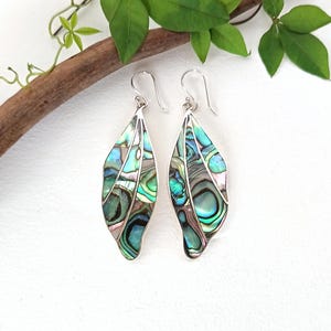 Può includere: Un paio di orecchini pendenti con ciondoli a forma di foglia allungata. Ogni ciondolo è realizzato con segmenti di conchiglia di abalone iridescente nei toni del blu, verde e viola, separati da sottili linee di metallo argentato con ganci.