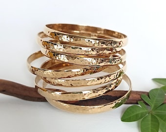 Puede incluir: Una pila de cinco brazaletes de metal de color dorado, cada uno con una superficie martillada y texturizada que refleja la luz. Los brazaletes circulares están dispuestos en una pila vertical, mostrando un acabado metálico uniforme y un perfil redondeado.