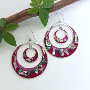 May include: A pair of silver hoop earrings with a magenta inlay and iridescent abalone shell accents. Each earring features a large outer hoop, a smaller inner hoop, and a central circle. The earrings are displayed on a white surface.