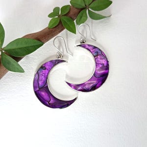 Può includere: Un paio di orecchini a forma di mezzaluna con un intarsio in conchiglia iridescente viola vibrante, incastonati in una montatura in metallo color argento lucido. Ogni orecchino pende da un semplice gancio argentato e presenta una superficie liscia.
