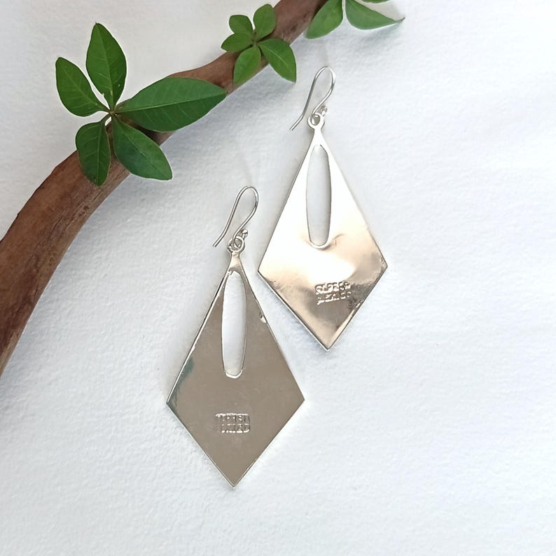 May include: A pair of polished silver dangle earrings featuring a diamond-shaped silhouette with a central vertical cutout. Each earring includes a curved hook wire and is stamped with the text 925 Mexico on the lower portion of the metal surface.