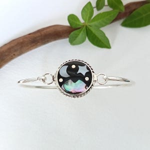 以下が含まれることがあります： 丸い黒いセンターピースが付いたシルバーのバングルブレスレット。 センターピースは三日月と星のデザインで、虹色のあわびの貝殻のアクセントが付いています。 ブレスレットは繊細なジュエリーです。