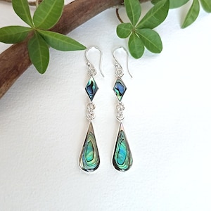 May include: A pair of silver drop earrings featuring iridescent abalone shell inlays. Each earring consists of a small diamond-shaped piece connected by a silver link to a larger teardrop-shaped pendant, suspended from a curved silver ear wire.