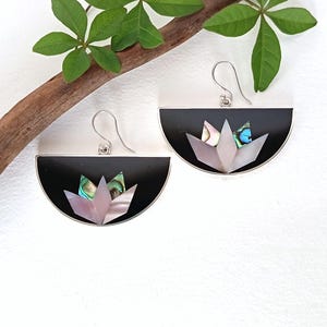 Peut inclure: Une paire de boucles d'oreilles semi-circulaires avec une base noire et un motif floral incrusté. L'incrustation se compose de fragments de nacre d'ormeau irisés et de pétales en nacre rose pâle. Chaque boucle pend à un crochet en métal argenté.