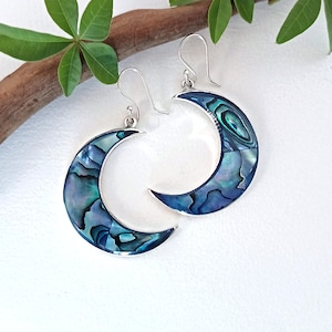 May include: A pair of crescent moon-shaped earrings featuring iridescent abalone shell inlay set in silver-toned metal frames. Each earring hangs from a simple silver-toned hook wire, showcasing a mosaic of blue, green, and purple shell segments.