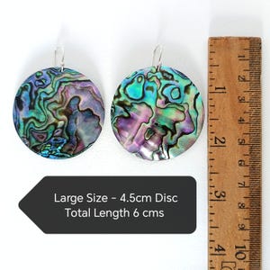 May include: Two iridescent abalone shell earrings with silver hooks. Each earring is a large, round disc, approximately 1.75 inches in diameter, with a total length of 2.36 inches. The earrings display a mix of blues, greens, and purples.