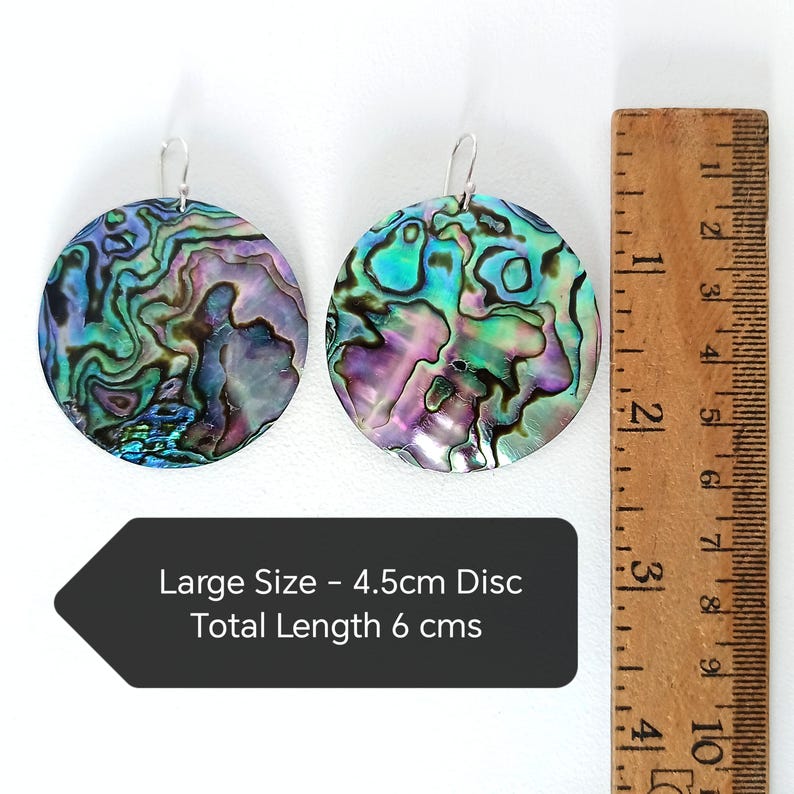 May include: Two iridescent abalone shell earrings with silver hooks. Each earring is a large, round disc, approximately 1.75 inches in diameter, with a total length of 2.36 inches. The earrings display a mix of blues, greens, and purples.