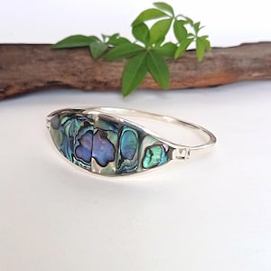 Op de afbeelding: Een zilverkleurige scharnierende armband met een centrale gebogen inleg van iriserende abalone-schelp. De schelp vertoont een levendige mix van blauwe, groene en paarse tinten met een gepolijste afwerking, gevat in een glad, reflecterend metalen frame.