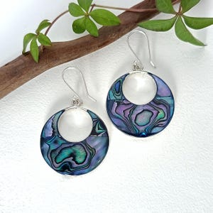 May include: Silver-tone hoop earrings with iridescent abalone shell in blue, green, and purple hues. Each earring has a circular design with a central white circle and a hook fastening. The earrings are presented on a white surface with a wooden branch and green leaves.