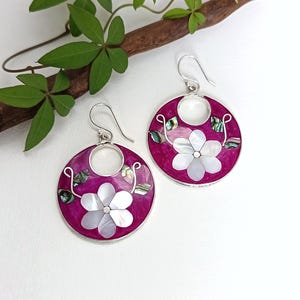 Peut inclure: Une paire de boucles d'oreilles rondes magenta avec un crochet argenté. Chaque boucle d'oreille présente un motif de fleur blanche au centre, entouré d'accents irisés et d'une bordure argentée. Les boucles d'oreilles sont posées sur un fond blanc.