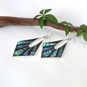 May include: A pair of dangle earrings featuring a geometric diamond shape with a black frame and inlaid iridescent abalone shell segments. The earrings include silver-toned metal hooks for attachment and display a vibrant blue and green color palette.