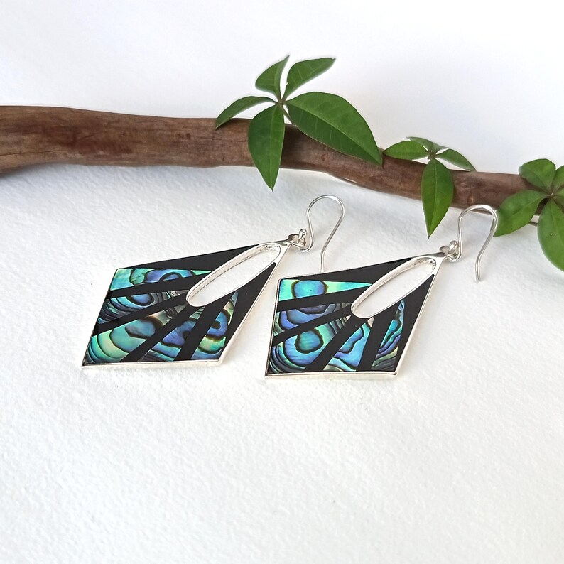 May include: A pair of dangle earrings featuring a geometric diamond shape with a black frame and inlaid iridescent abalone shell segments. The earrings include silver-toned metal hooks for attachment and display a vibrant blue and green color palette.