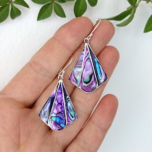 Peut inclure: Boucles d'oreilles en argent avec un design géométrique triangulaire. Les boucles d'oreilles présentent des coquillages d'ormeau irisés dans des tons de violet, de bleu et de vert, sertis dans un cadre argenté. Les boucles d'oreilles sont présentées sur un fond neutre.