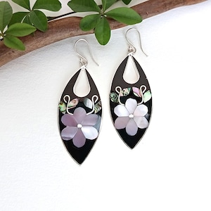 May include: A pair of teardrop-shaped earrings featuring a black base with inlaid iridescent shell floral motifs. Each earring has a silver-toned hook and a central flower design composed of light purple and white shell petals with green accents.