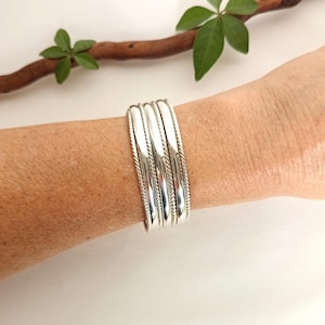 May include: A set of four silver-toned cuff bracelets worn on a wrist. Each bracelet features a smooth, polished central band flanked by two thinner, twisted rope-style silver wires, creating a layered and textured appearance on the arm.