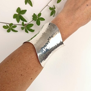 Op de afbeelding: Een brede, zilverkleurige manchetarmband met een gehamerd, getextureerd oppervlak dat een reflecterende, onregelmatige afwerking op het metaal creëert. De armband heeft een gebogen, open ontwerp dat om de pols valt en een gepolijste glans vertoont.