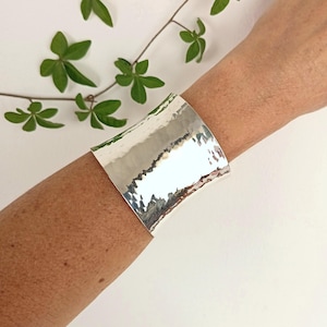 Op de afbeelding: Een brede, gepolijste zilveren manchetarmband met een gehamerde textuur die een onregelmatig, reflecterend oppervlak creëert. De metalen band wikkelt zich om de pols en toont een heldere, metallic afwerking met duidelijke inkepingen.