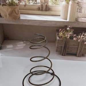 May include: A rusty metal spring with a circular base and a coiled upper section. The spring stands upright on a white surface, with a blurred background featuring a window and floral arrangements. The spring's design suggests a vintage or industrial aesthetic.