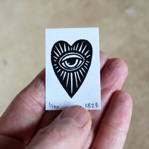 May include: Black and white print of an eye inside a heart with radiating lines. The print is numbered 1/100 and 0323.
