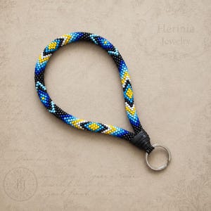 May include: A wristlet keychain crafted from small glass beads arranged in a geometric pattern of black, blue, yellow, and white. The beaded loop is secured with a black leather-like connector attached to a circular silver-toned metal key ring.