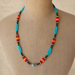 Op de afbeelding: Een kralenketting met afwisselende segmenten van turkooisblauwe, rode, oranje en gele kraaltjes, gescheiden door kleine donkerbruine tussenkralen. Het ontwerp bevat een enkele lichtblauwe gespikkelde kraal in het midden onderaan.